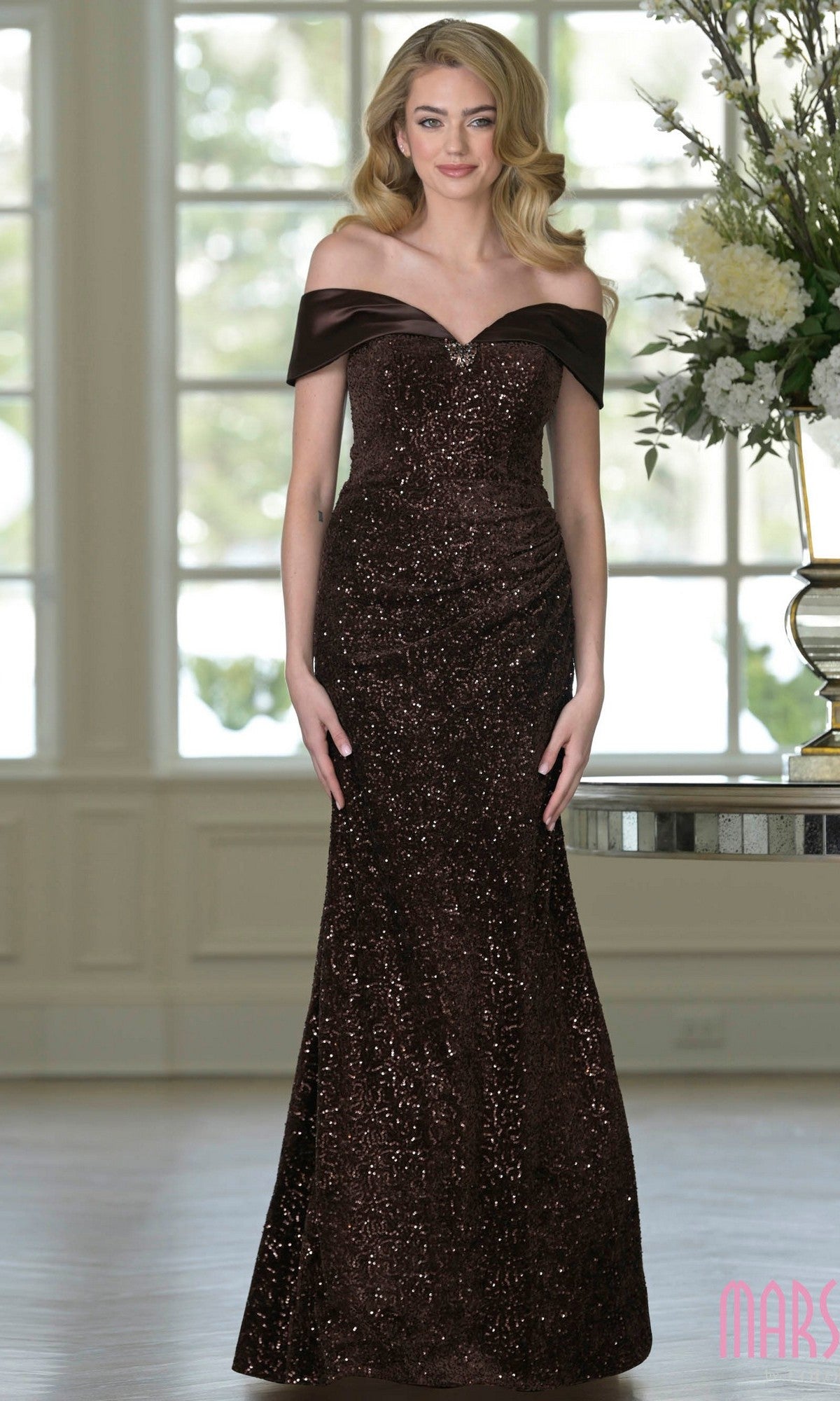 A woman poses indoors by large windows and floral arrangements, wearing the Marsoni M349 long formal dress in chocolate—a chic off-the-shoulder evening gown embellished with sequins. She has long, wavy hair.